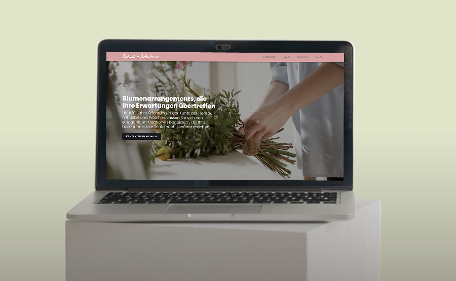 Laptop on a white pedestal displaying a floral arrangement website with pink header and German text.